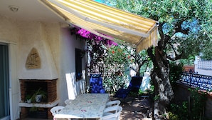 Une terrasse avec une table et des chaises, une cheminée en briques et une pergola surmontée d’une marquise à rayures.