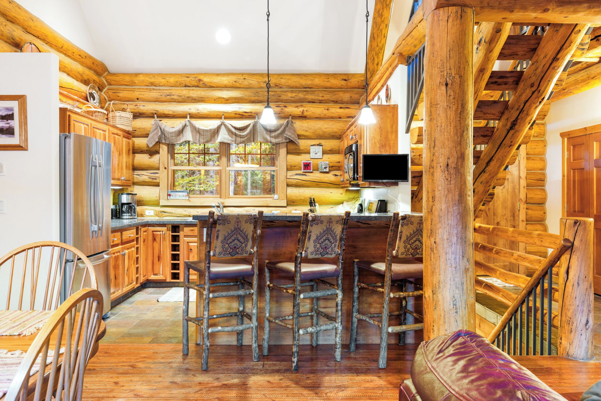 A rustic log cabin interior with a wooden dining table, bar stools, a kitchen area, and a staircase.