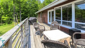 A deck with wicker furniture and a barbecue grill, overlooking a wooded area. - Mountain Haus: 4 br, a/c, firepit, free ski shuttle! (Stowe)
