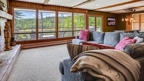 A cozy living room with a large window overlooking a scenic landscape, a fireplace, and a dining area with a view. - Mountain Haus: 4 br, a/c, firepit, free ski shuttle! (Stowe)