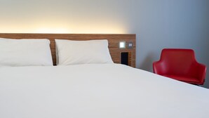 Habitació d'hotel amb un capçal de llit de fusta, dos coixins blancs, una cadira vermella i una taula de nit amb una làmpada.