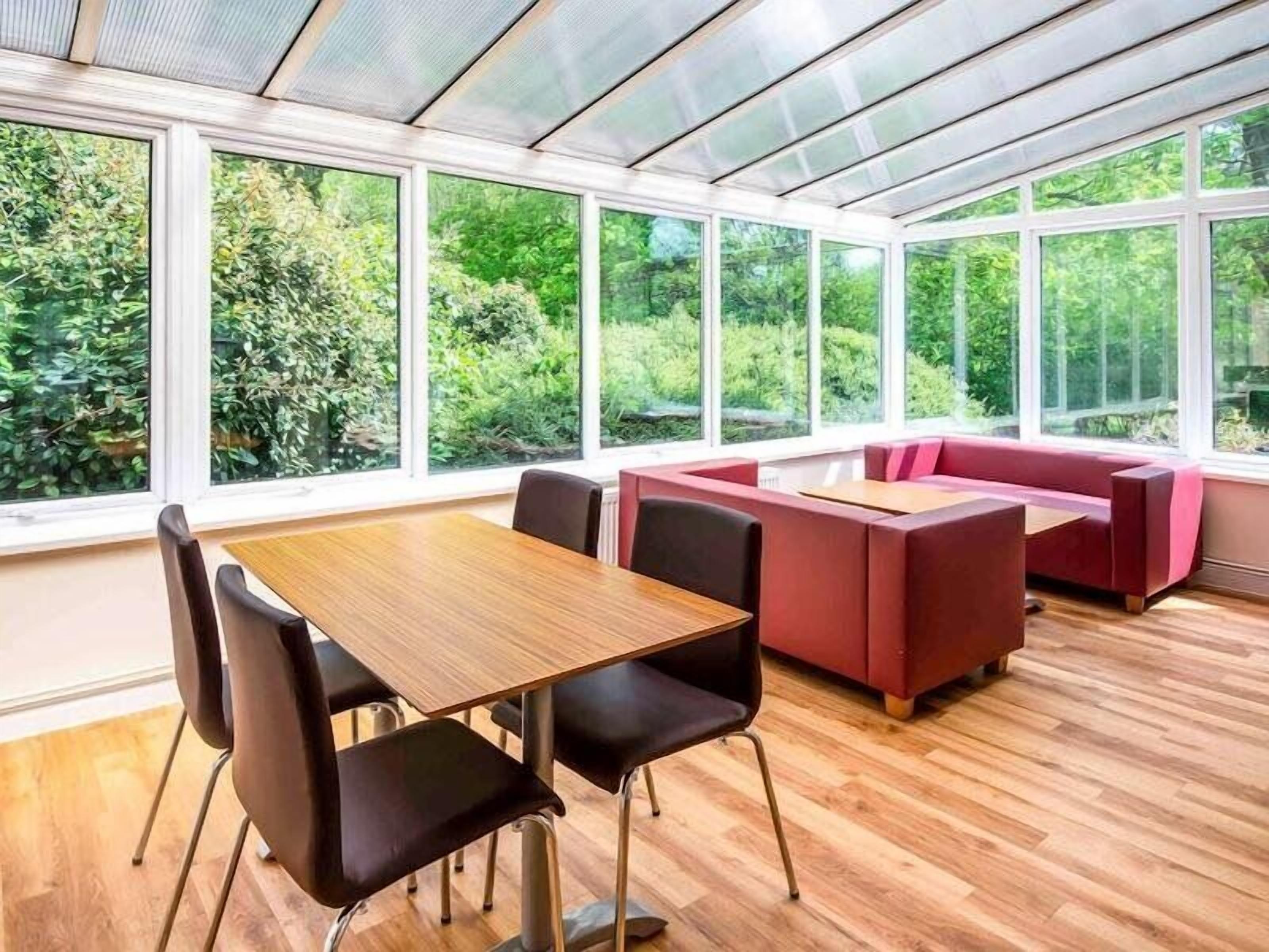 Une véranda avec une table et des chaises en bois, des sièges rouges et de grandes fenêtres donnant sur un paysage verdoyant.