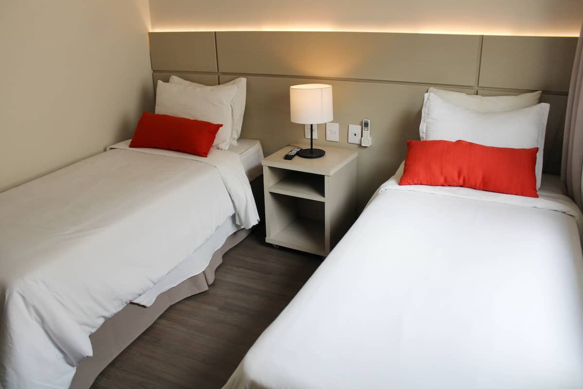 A hotel room with two beds, each with white bedding and red pillows. There is a nightstand between the beds with a lamp and a remote control.