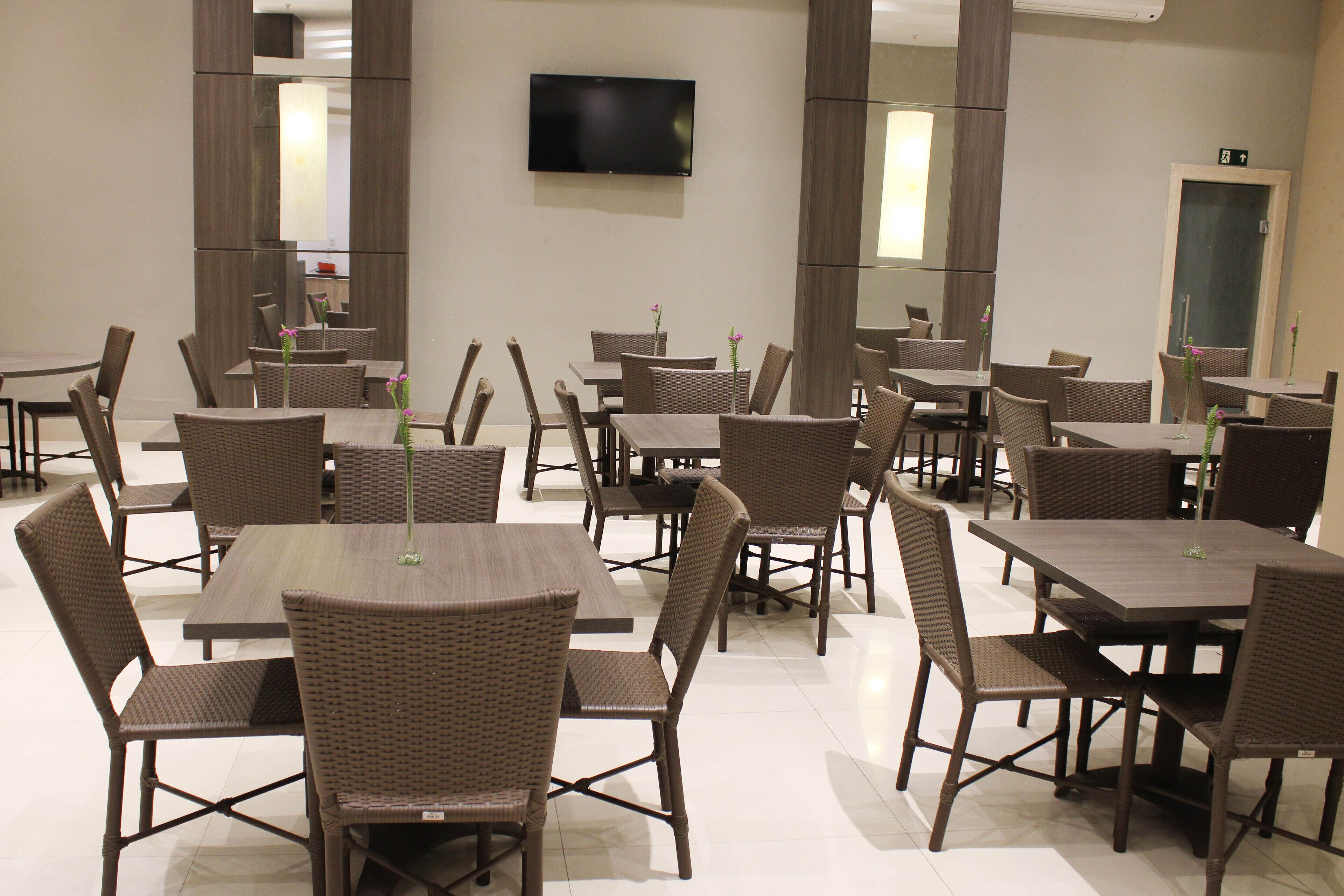 A hotel dining area with multiple tables and chairs, a flat-screen TV mounted on the wall, and a door with an emergency exit sign.
