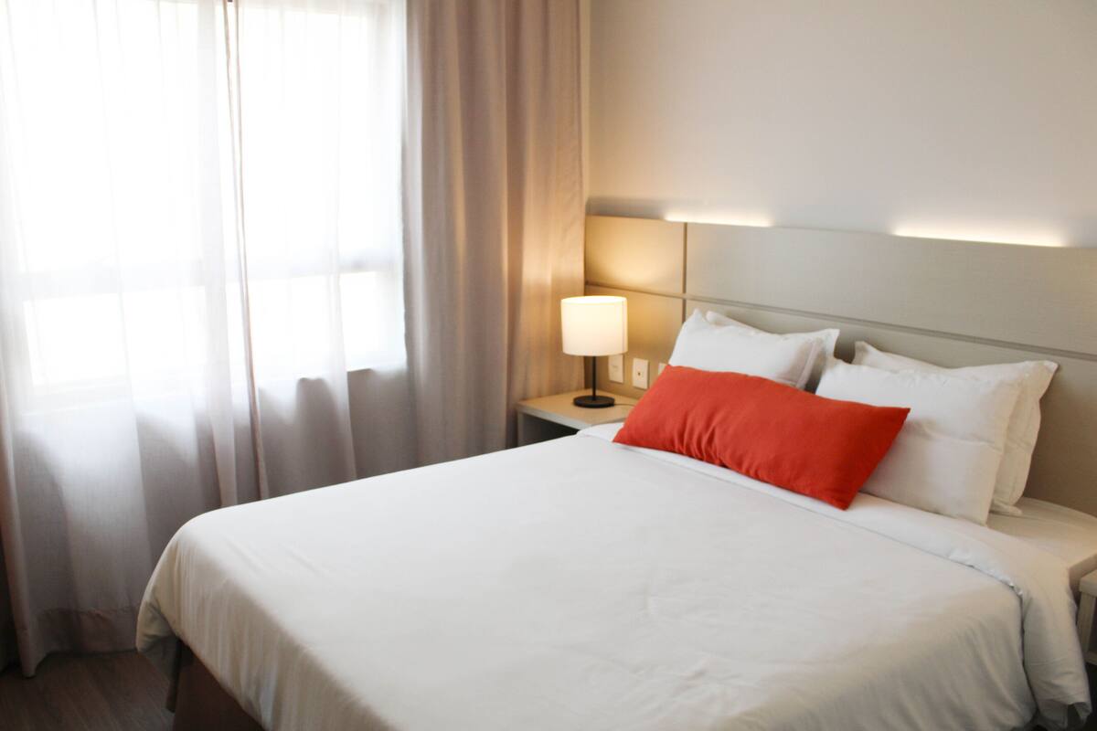 A hotel room with a bed, a nightstand, a lamp, and a window with curtains.