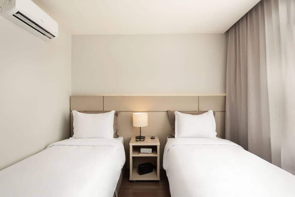 A hotel room with two beds, a nightstand with a lamp, and an air conditioning unit.