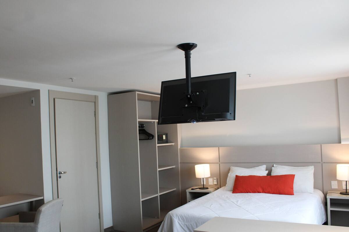 A hotel room with a bed, a television mounted on the ceiling, a desk, and a chair.