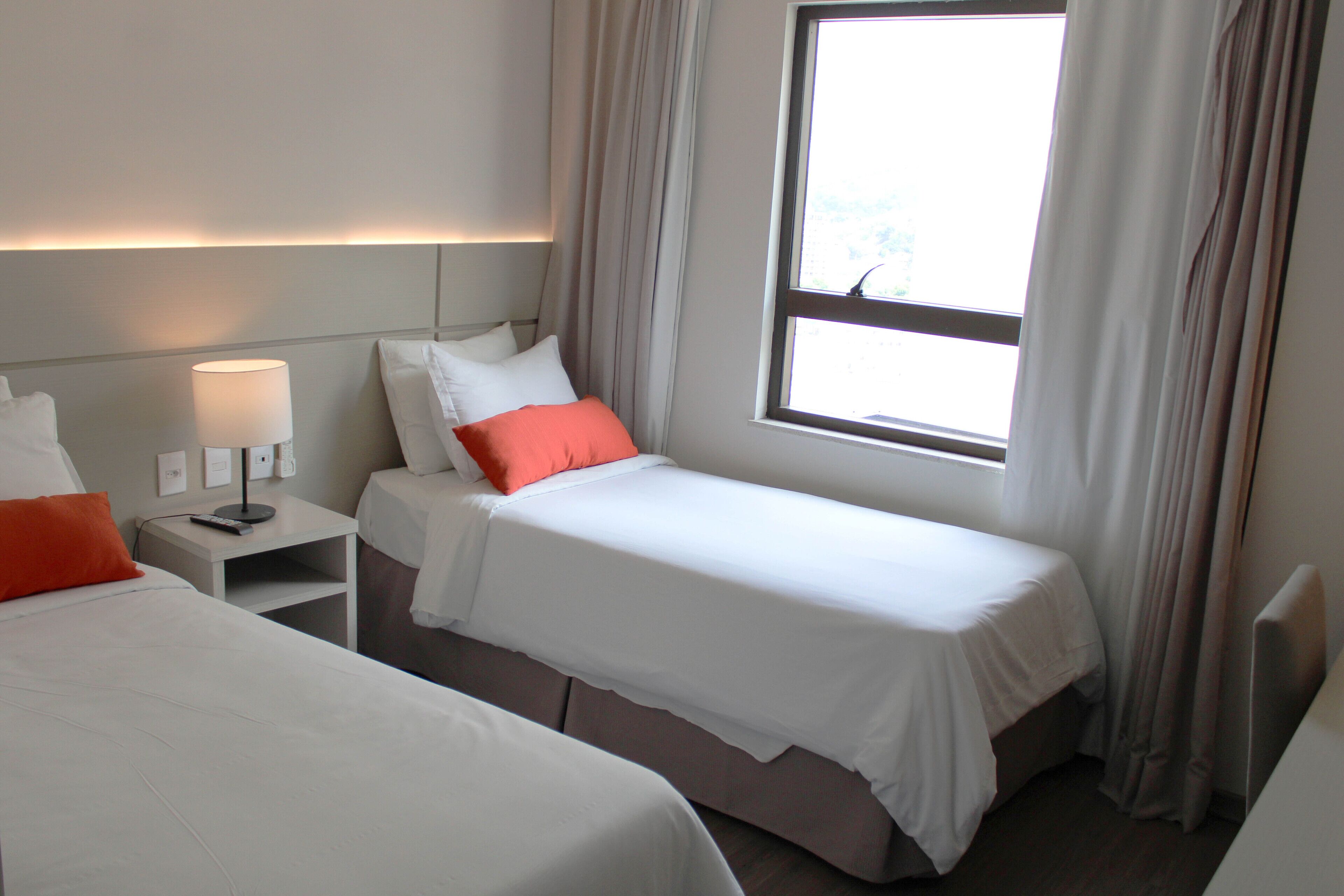 A hotel room with two single beds, a nightstand with a lamp, and a window with curtains.