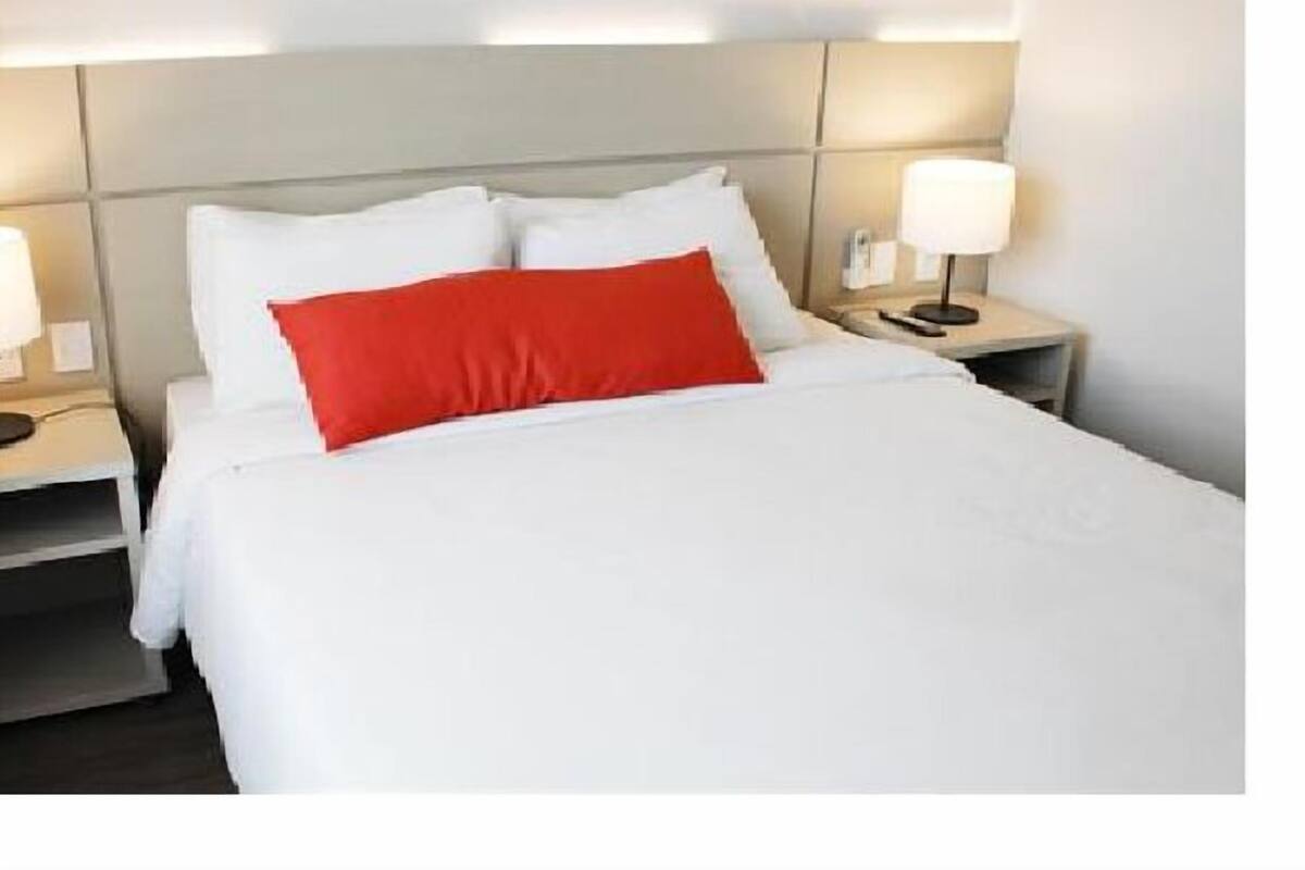 A hotel room with a bed, white bedding, a red accent pillow, and bedside lamps.