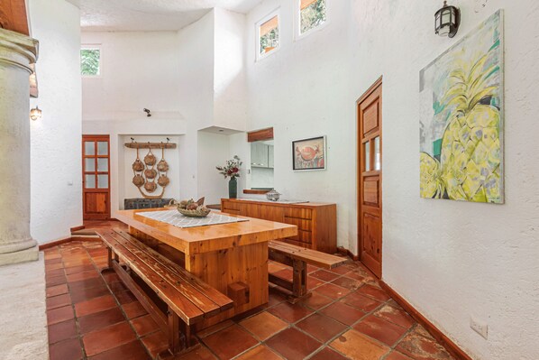 Una cocina amplia con una mesa de madera y bancos, paredes blancas y suelo de baldosas.