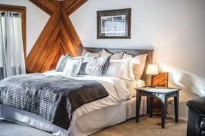 A bedroom with a bed, pillows, a bedside table with a lamp, a framed picture on the wall, and a window with curtains.