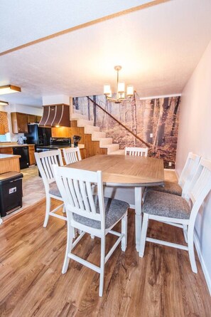 A dining area with a wooden table and white chairs, a kitchen in the background, and a staircase leading to another level. - Comfy house: 3 king beds, hot tub, attached garage (Red Lodge)