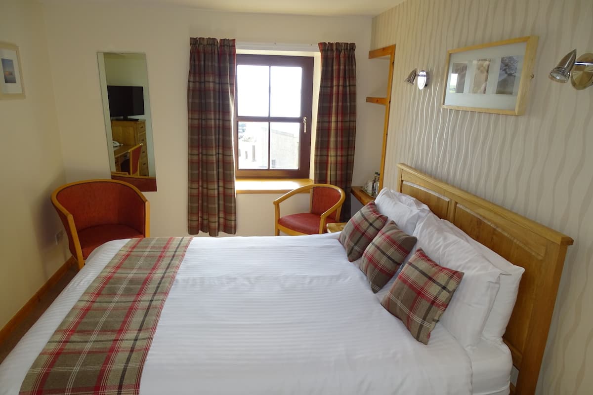 A hotel room with a large bed, two chairs, a wooden headboard, a window with curtains, a mirror, and a wall-mounted picture frame.