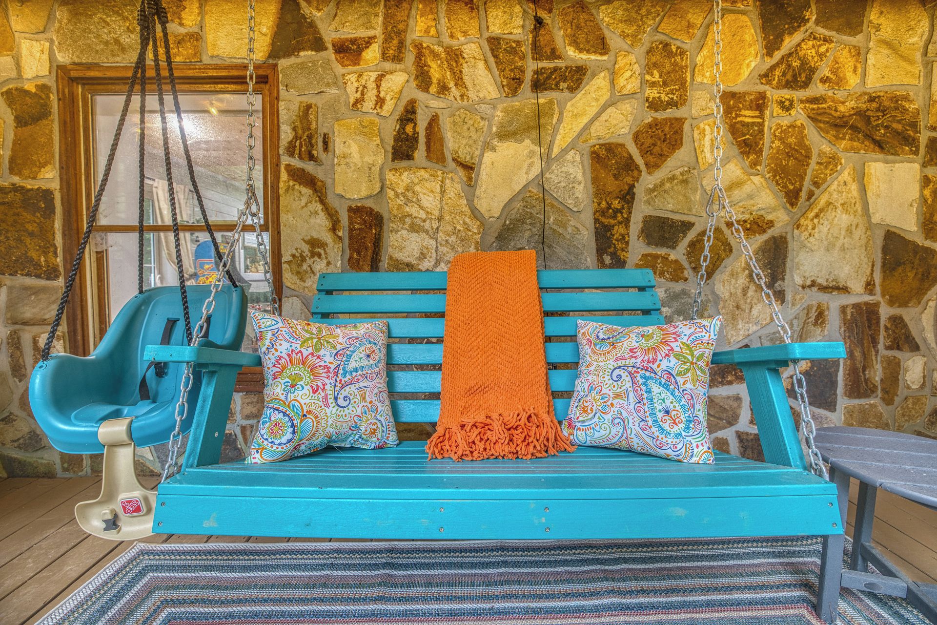 A turquoise swing set with a woven rug, colorful cushions, and a stone wall backdrop.