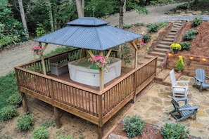 A wooden deck with a hot tub, surrounded by a stone patio and outdoor furniture.