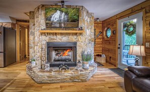 A stone fireplace with a fire, surrounded by stone walls and a wooden ceiling. - Bearfoot Bliss-Bear Hunt • Arcade • Hot Tub- Fire Pit- Adventure Cabin-Sleeps 12 (Blairsville)