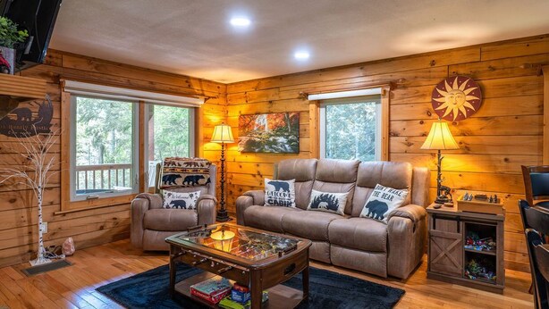 Ein gemütliches Blockhaus-Interieur mit einer Couch, Sesseln und einem Couchtisch. Zwei Fenster, eine Holzwand und eine Lampe auf einem Beistelltisch sind zu sehen.
