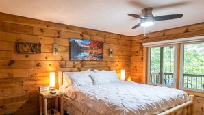 Ein Schlafzimmer in einer Holzhütte mit Bett, Nachttisch, Lampe, Deckenventilator und einem Fenster mit Blick auf Grünflächen.