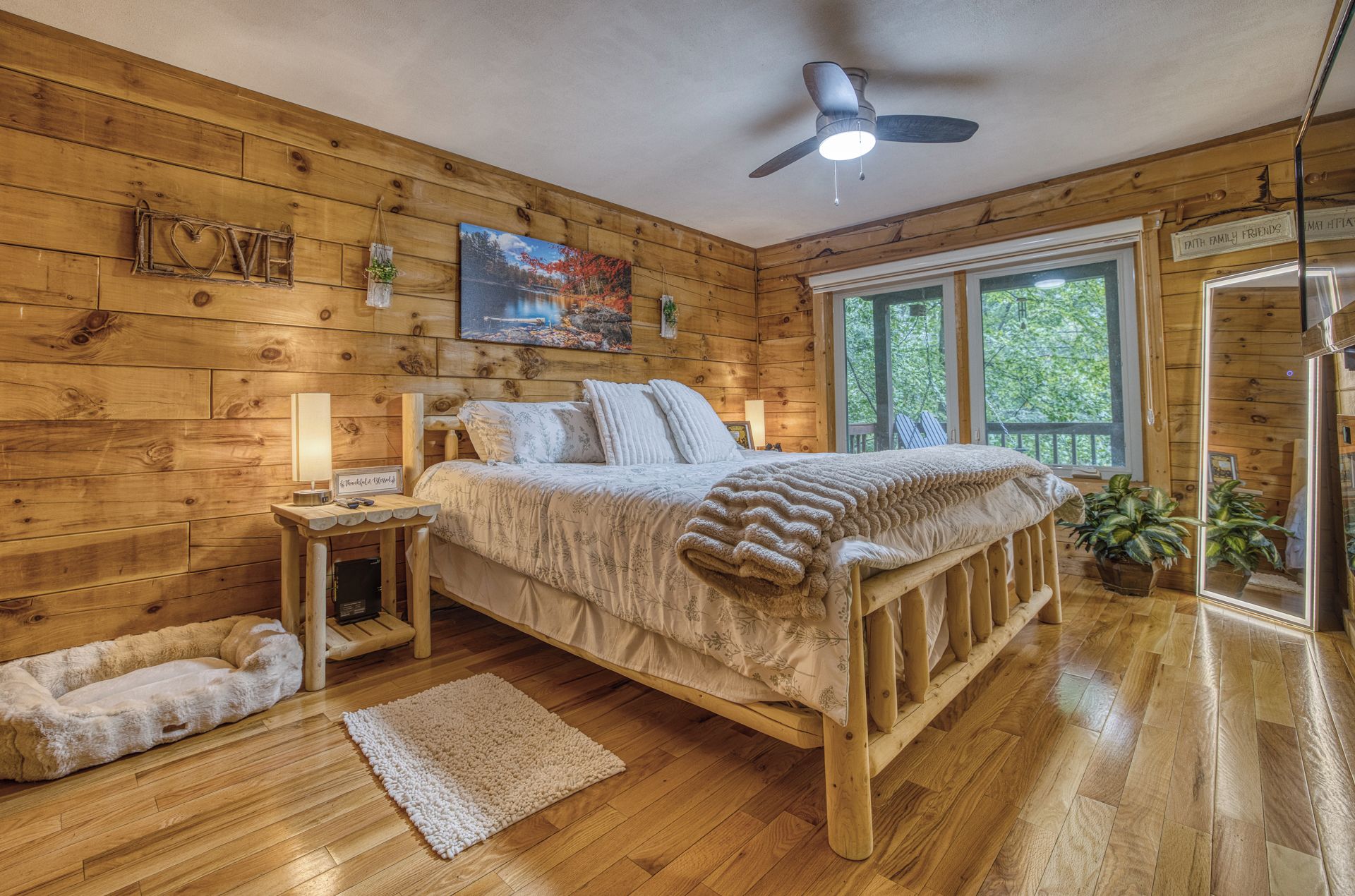 Une chambre confortable aménagée dans un chalet en bois, avec un lit, une table de chevet, un coussin moelleux pour chien, un tapis et une grande fenêtre donnant sur la forêt.