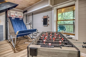 A room with a foosball table, a basketball hoop, and a window with a view of trees.