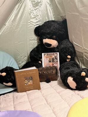 A black teddy bear sitting on a bed with a book titled 'Explorer Scrapbook' and a small wooden chest.