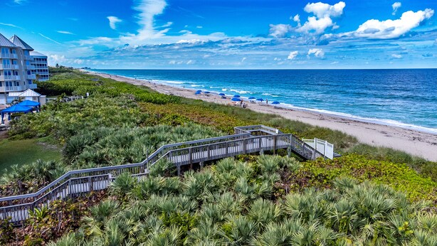 Un hôtel situé sur la côte, avec une promenade en bois menant à la plage, le tout entouré d’une végétation luxuriante.
