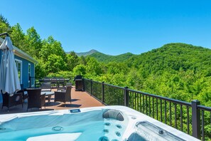 A hot tub on a deck with outdoor furniture, overlooking a forested mountain.