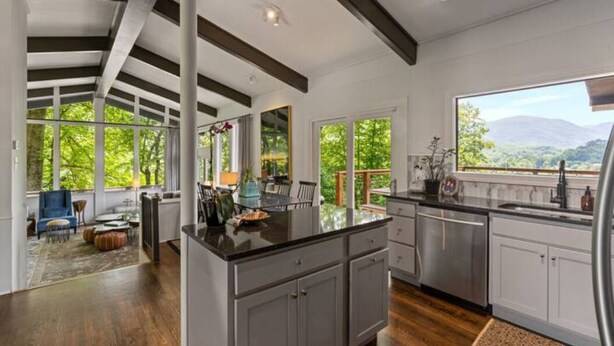 A spacious kitchen with a large island, dark wooden floors, and a view of greenery through large windows. - Mid-century home with panoramic views and hot tub! (Maggie Valley)