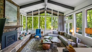A spacious living room with a stone fireplace, large windows, and a view of greenery. - Mid-century home with panoramic views and hot tub! (Maggie Valley)