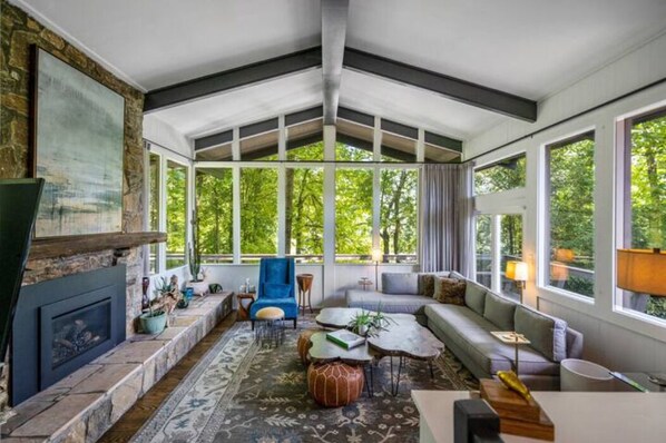 A spacious living room with a stone fireplace, large windows, and a view of greenery.