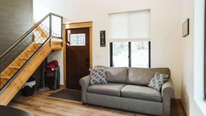 Un interior moderno con una escalera de madera, un sofá gris con cojines estampados y un ventanal con persianas.