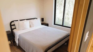 A bedroom with a bed, white bedding, a nightstand, a lamp, and a window with a view of trees. - Lynx Cabin @ Keystone Kozy (Soldotna)