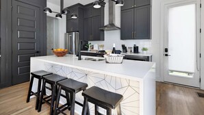 Una cocina moderna con armarios oscuros, una isla blanca con taburetes de bar, electrodomésticos de acero inoxidable e iluminación colgante.