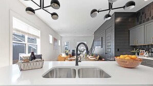 Una cocina moderna con encimera blanca, pileta doble y muebles oscuros. Se ve un comedor con mesa y sillas al fondo.