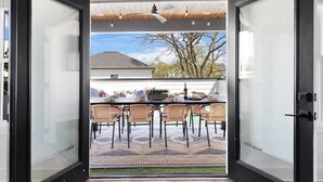 Puerta corredera de vidrio abierta que da a un patio exterior con una mesa y sillas, un ventilador de techo y vistas a una casa y árboles.
