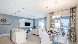Un salon moderne doté d’un îlot de cuisine, d’un coin repas avec des chaises blanches, d’un canapé et d’une grande fenêtre agrémentée de rideaux à motifs.