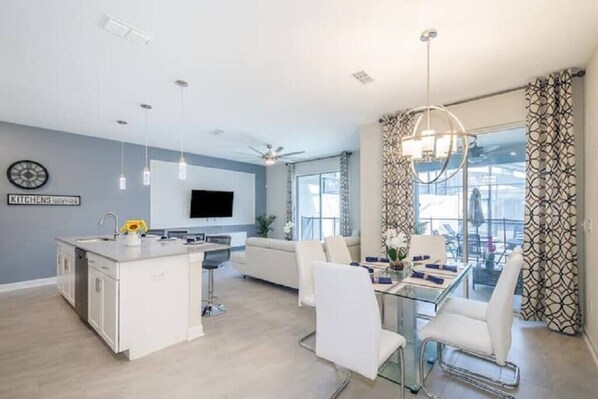 A modern living room with a kitchen island, a dining area with white chairs, a sofa, and a large window with patterned curtains.