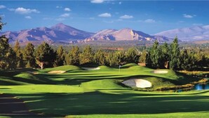 A golf course with bunkers and a flagstick, surrounded by trees and mountains.