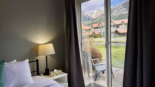 A hotel room with a bed, a bedside table with a lamp, a tissue box, and a view of a mountain landscape through a large window. - Cozy Rocky Mountain Chalet | Pet Friendly (Canmore)