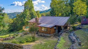 A rustic cabin with a stone foundation, a wooden exterior, and a metal roof, surrounded by lush greenery and trees. - Best View! Beautiful Ozarks cabin ON rim of AR Grand Canyon! Many new updates (Jasper)