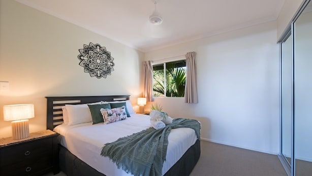 Une chambre à coucher comprenant un lit, des tables de chevet, un élément de décoration murale, une fenêtre avec des rideaux et une vue sur la verdure à l’extérieur.