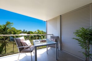 A balcony with a table and chairs, a view of greenery, and a closed sliding door. - Waterlily - One Bedroom Apartment (Airlie Beach)