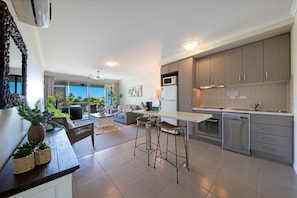 A modern kitchen with a dining area, bar stools, and a view of the outdoors. - Waterlily - One Bedroom Apartment (Airlie Beach)