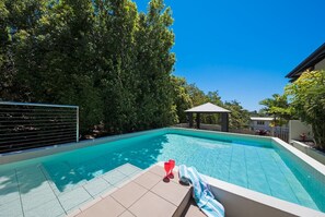 A swimming pool with a tiled deck, surrounded by trees and a gazebo. - Waterlily - One Bedroom Apartment (Airlie Beach)