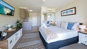 Quarto com cama grande, mesa de cabeceira, cômoda, espelho e televisão fixada na parede.