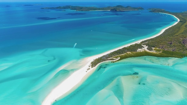 Vista aérea de uma área costeira com águas turquesa cristalinas, praias de areia branca e uma faixa estreita de vegetação separando a praia da água.