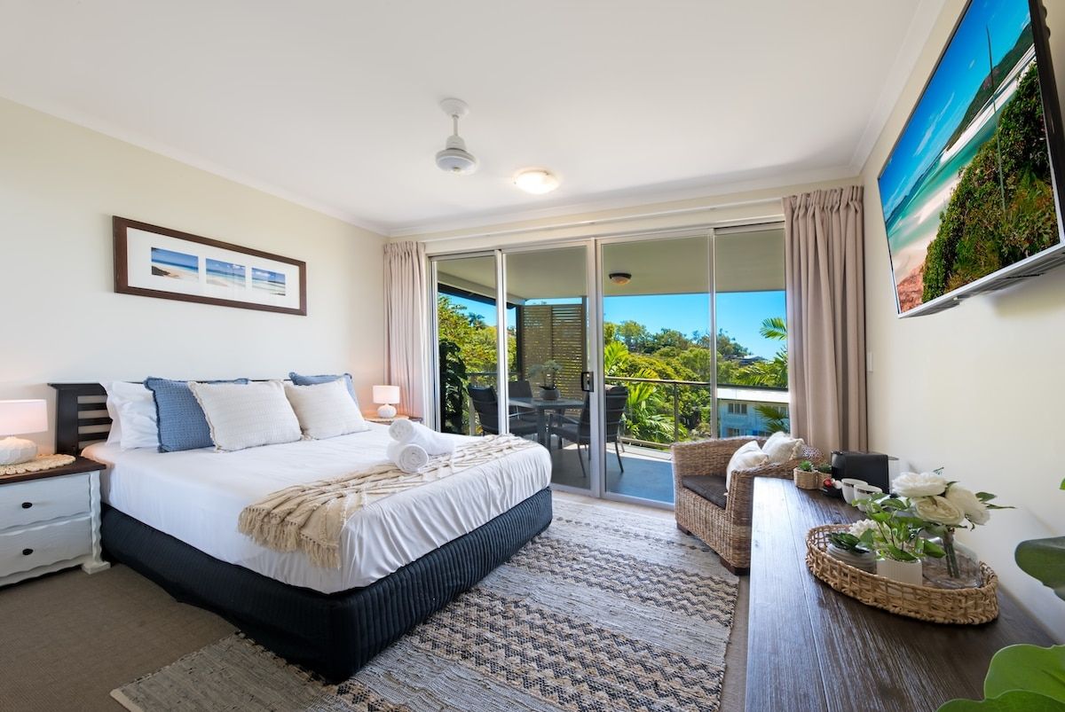 A bedroom with a bed, bedside tables, a nightstand, a large window, and a view of greenery outside.