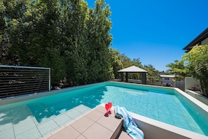 A swimming pool with a tiled deck, surrounded by trees and a gazebo. - Waterlily - Two Bedroom Apartment (Airlie Beach)