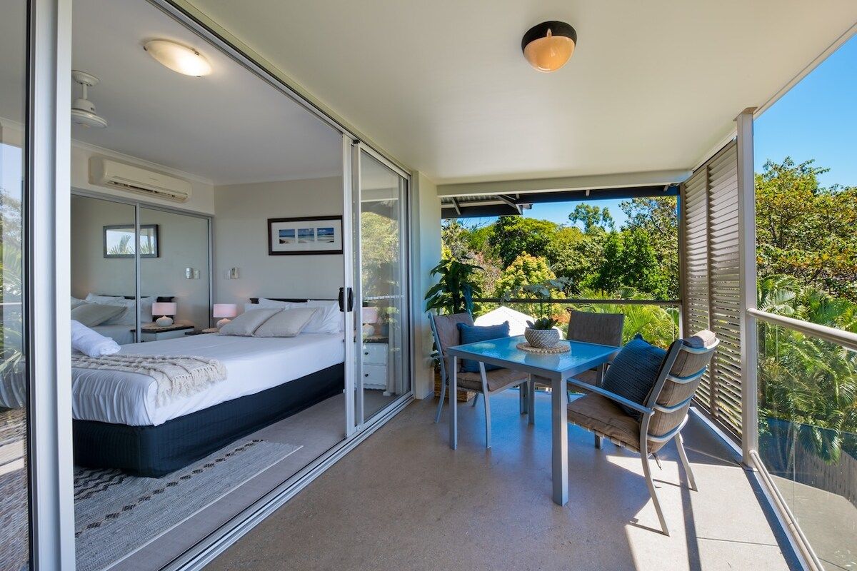 Een moderne slaapkamer met een glazen deur die leidt naar een balkon, een zithoek met buitenmeubilair en een helder uitzicht op de omliggende groene omgeving.