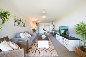 A modern living room with a grey sofa, a coffee table, and a TV on a white cabinet. There are plants and artwork on the walls. - Waterlily - Two Bedroom Apartment (Airlie Beach)